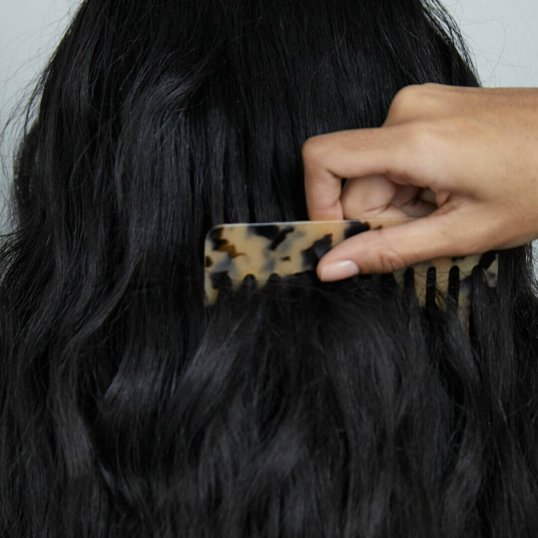 A brunette woman parting and combing her long, thick hair, demonstrating how to get thicker hair with proper care and styling techniques.